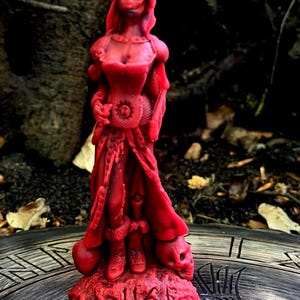 Könnte beinhalten: Eine rote Kerze in Form einer Frau in einem langen Kleid, mit einem Totenkopf am Fuß. Die Kerze hat detaillierte Elemente wie ein korsettartiges Oberteil und fließende Ärmel. Der Sockel hat geschnitzte Symbole. Die Kerze steht auf einer Holzoberfläche.