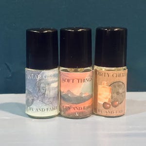 May include: Three small glass bottles with black caps and labels. The labels have different designs and text. The first bottle says "Star Girl" with a drawing of a woman's face. The second bottle says "Soft Things" with a drawing of mountains. The third bottle says "Dirty Cherry" with a drawing of cherries and a clock.