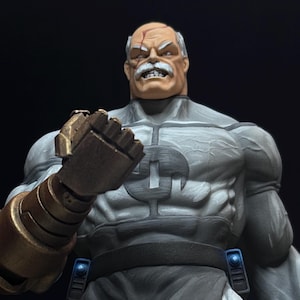 Puede incluir: Figura detallada de un personaje musculoso con expresión severa. La figura es principalmente gris con bigote blanco y cicatriz en la frente. El brazo derecho presenta un gran guantelete metálico con detalles azules.