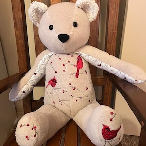 Puede incluir: Un oso de peluche beige con orejas texturizadas, ojos negros y nariz negra. El torso y los pies presentan un patr&oacute;n bordado de cardenales rojos posados en ramas delgadas con peque&ntilde;as bayas rojas. El interior de los brazos tiene un estampado floral.
