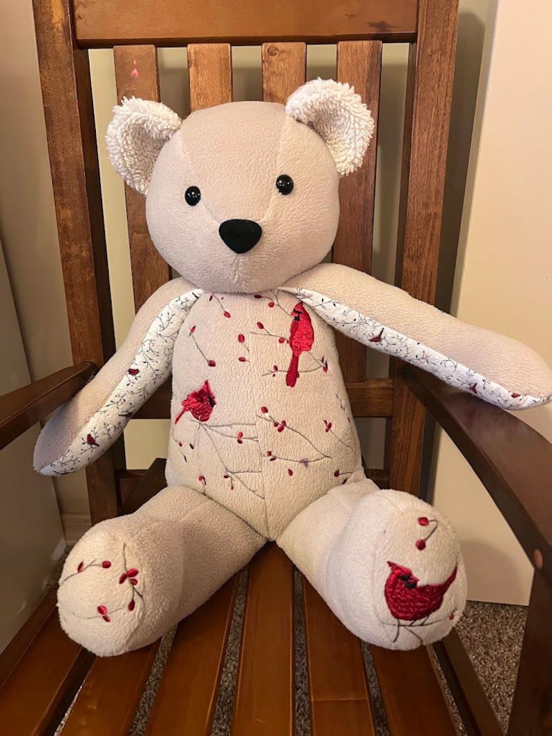 Puede incluir: Un oso de peluche beige con orejas texturizadas, ojos negros y nariz negra. El torso y los pies presentan un patr&oacute;n bordado de cardenales rojos posados en ramas delgadas con peque&ntilde;as bayas rojas. El interior de los brazos tiene un estampado floral.