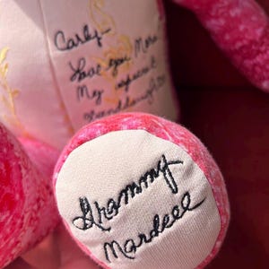 Puede incluir: Un oso de peluche de tela con estampado rosa que presenta texto negro bordado en cursiva sobre su pecho y planta del pie de color crema. El pecho dice Carly Love you More My sweet Granddaughter y la planta del pie tiene bordado Grammy Mardeee.