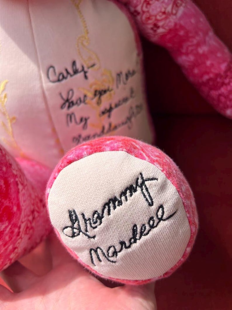 Puede incluir: Un oso de peluche de tela con estampado rosa que presenta texto negro bordado en cursiva sobre su pecho y planta del pie de color crema. El pecho dice Carly Love you More My sweet Granddaughter y la planta del pie tiene bordado Grammy Mardeee.