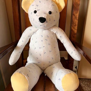 Puede incluir: Un oso de peluche hecho de tela blanca con un patr&oacute;n repetitivo de peque&ntilde;as anclas azul oscuro y estrellas gris claro. El oso presenta tela amarilla s&oacute;lida en el interior de las orejas y las plantas de los pies, con ojos de bot&oacute;n y nariz negros.