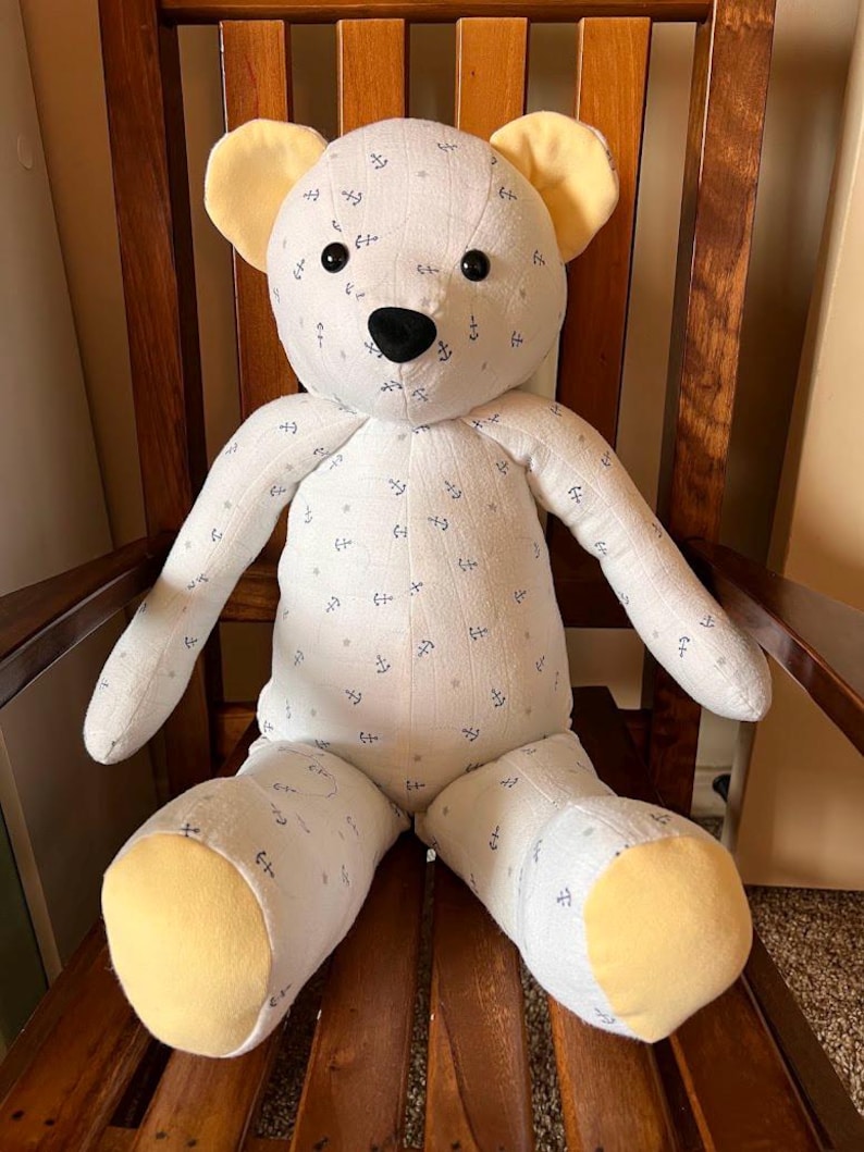 Puede incluir: Un oso de peluche hecho de tela blanca con un patr&oacute;n repetitivo de peque&ntilde;as anclas azul oscuro y estrellas gris claro. El oso presenta tela amarilla s&oacute;lida en el interior de las orejas y las plantas de los pies, con ojos de bot&oacute;n y nariz negros.