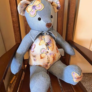 Puede incluir: Un oso de peluche hecho de tela de mezclilla azul. El oso presenta un panel en el pecho de color crema decorado con un patr&oacute;n de mariposas coloridas, detalles de mariposas a juego en las orejas y un pie, una nariz marr&oacute;n y ojos negros.