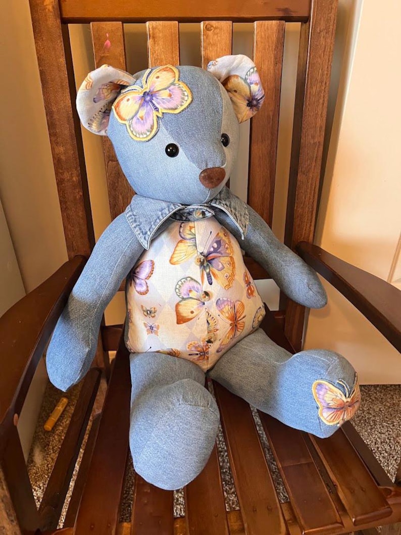 Puede incluir: Un oso de peluche hecho de tela de mezclilla azul. El oso presenta un panel en el pecho de color crema decorado con un patr&oacute;n de mariposas coloridas, detalles de mariposas a juego en las orejas y un pie, una nariz marr&oacute;n y ojos negros.