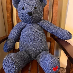 Puede incluir: Un oso de peluche tejido en color azul con una superficie texturizada, que presenta dos ojos negros redondos y una nariz negra cosida. Un peque&ntilde;o coraz&oacute;n rojo brillante est&aacute; bordado en la parte inferior de la pierna derecha del oso.