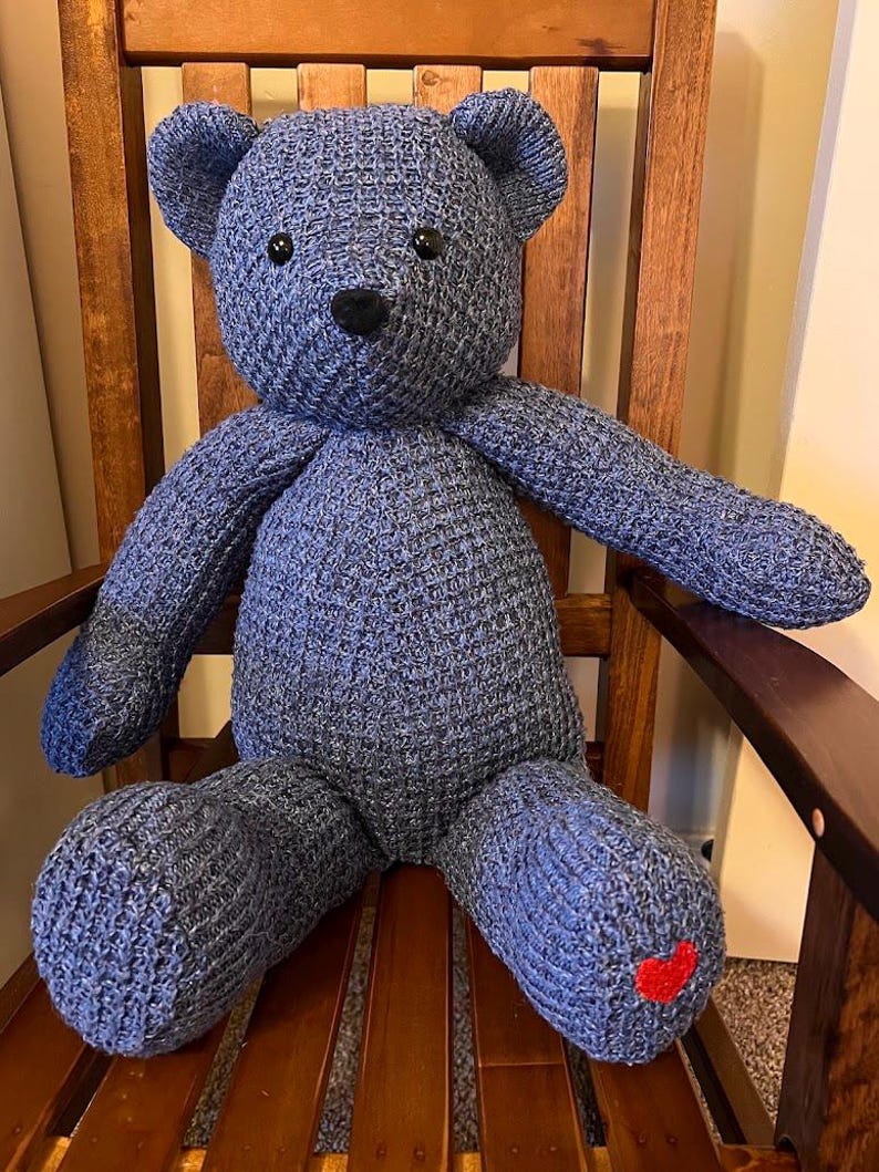 Puede incluir: Un oso de peluche tejido en color azul con una superficie texturizada, que presenta dos ojos negros redondos y una nariz negra cosida. Un peque&ntilde;o coraz&oacute;n rojo brillante est&aacute; bordado en la parte inferior de la pierna derecha del oso.