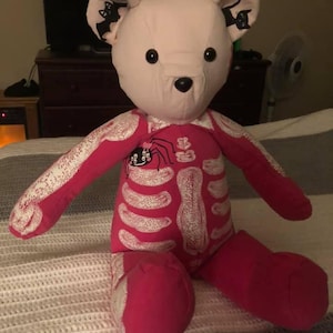 Puede incluir: Un oso de peluche con cabeza de color rosa claro y cuerpo de color rosa brillante con un estampado de esqueleto blanco. El oso tiene ojos de bot&oacute;n negros y una nariz negra. Las orejas internas est&aacute;n decoradas con siluetas negras de un murci&eacute;lago y un gato. Un peque&ntilde;o gr&aacute;fico de ara&ntilde;a con un lazo rosa est&aacute; impreso en la parte superior del pecho.