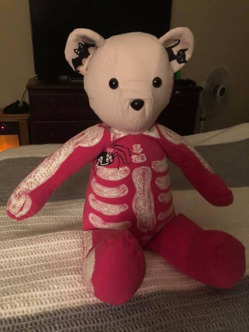 Puede incluir: Un oso de peluche con cabeza de color rosa claro y cuerpo de color rosa brillante con un estampado de esqueleto blanco. El oso tiene ojos de bot&oacute;n negros y una nariz negra. Las orejas internas est&aacute;n decoradas con siluetas negras de un murci&eacute;lago y un gato. Un peque&ntilde;o gr&aacute;fico de ara&ntilde;a con un lazo rosa est&aacute; impreso en la parte superior del pecho.