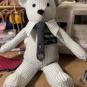 Puede incluir: Un oso de peluche hecho de tela a rayas azul claro y blanco. Lleva una corbata azul oscuro con lunares, un broche met&aacute;lico y una etiqueta negra con el nombre OSCAR D BAILEY CAPT. Los pies tienen bordadas las palabras Always with Dad.