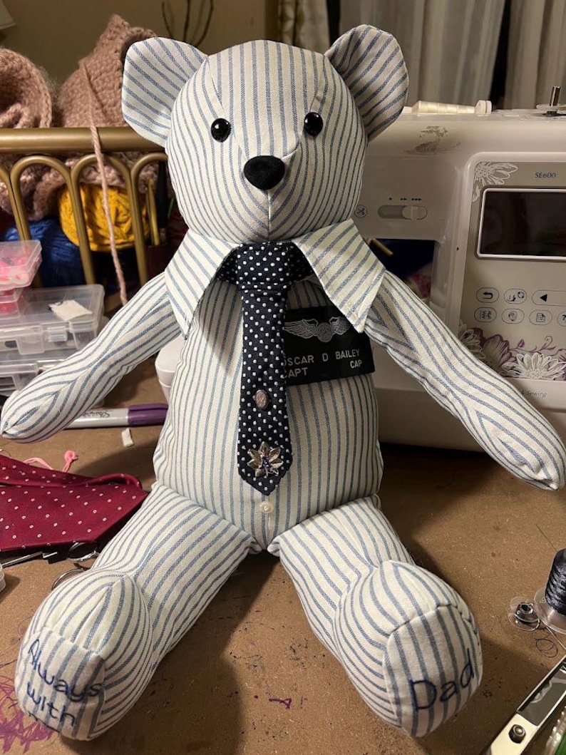Puede incluir: Un oso de peluche hecho de tela a rayas azul claro y blanco. Lleva una corbata azul oscuro con lunares, un broche met&aacute;lico y una etiqueta negra con el nombre OSCAR D BAILEY CAPT. Los pies tienen bordadas las palabras Always with Dad.