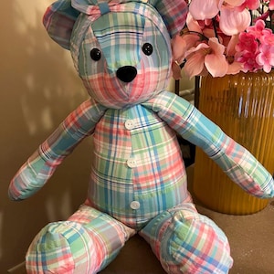 Puede incluir: Un oso de peluche hecho de tela a cuadros en azul claro, rosa y blanco. El oso presenta una diadema a juego atada con un lazo en la cabeza, dos ojos de bot&oacute;n negros, una nariz negra bordada y tres botones blancos en la parte frontal del torso.