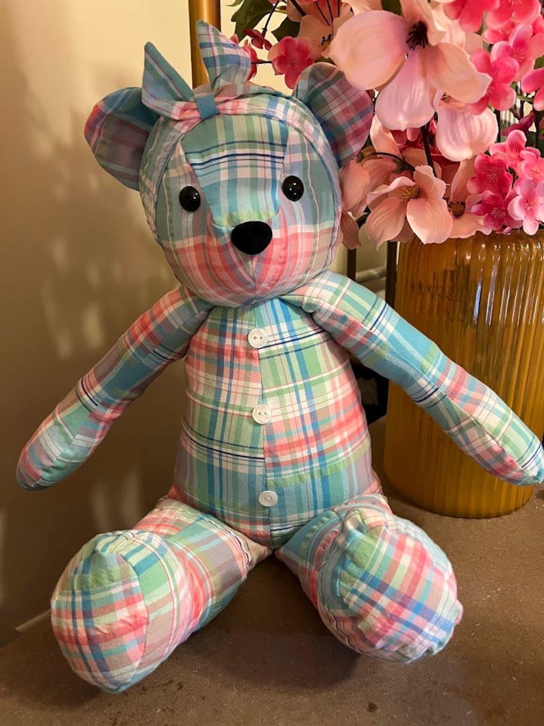 Puede incluir: Un oso de peluche hecho de tela a cuadros en azul claro, rosa y blanco. El oso presenta una diadema a juego atada con un lazo en la cabeza, dos ojos de bot&oacute;n negros, una nariz negra bordada y tres botones blancos en la parte frontal del torso.