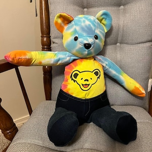Puede incluir: Un oso de peluche con cabeza y brazos te&ntilde;idos con efecto tie-dye en tonos azul, amarillo y naranja. El torso muestra un patr&oacute;n tie-dye amarillo con un gr&aacute;fico de cara de oso amarilla con contorno negro. La parte inferior es de mezclilla azul.