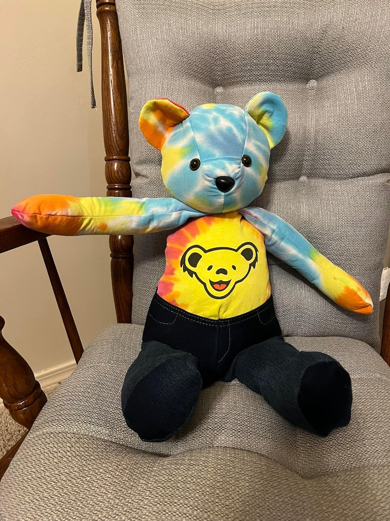 Puede incluir: Un oso de peluche con cabeza y brazos te&ntilde;idos con efecto tie-dye en tonos azul, amarillo y naranja. El torso muestra un patr&oacute;n tie-dye amarillo con un gr&aacute;fico de cara de oso amarilla con contorno negro. La parte inferior es de mezclilla azul.