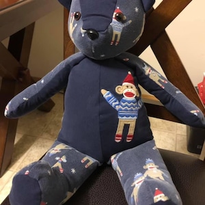 Puede incluir: Un oso de peluche hecho de tela azul oscuro con un patr&oacute;n repetitivo de monos de calcet&iacute;n con gorros rojos y su&eacute;teres azules. El oso tiene un panel de pecho azul s&oacute;lido con una aplicaci&oacute;n bordada de un mono de calcet&iacute;n.