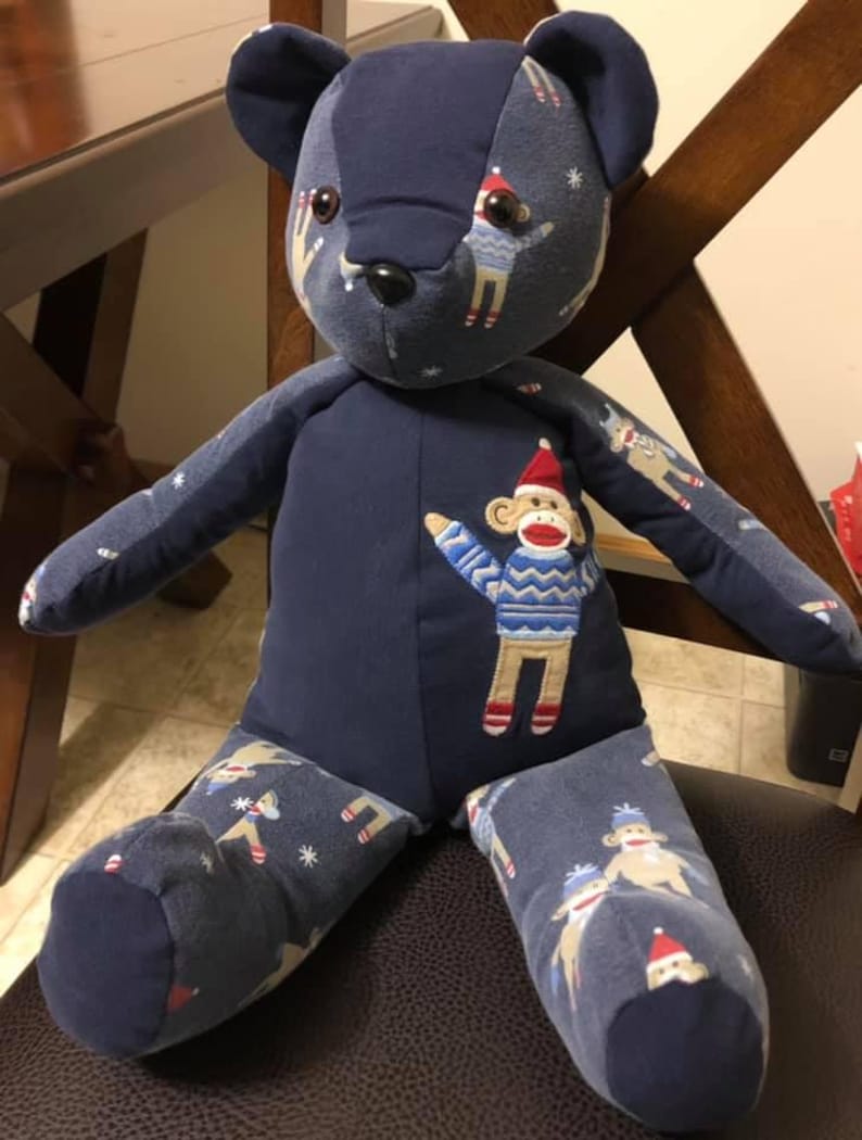 Puede incluir: Un oso de peluche hecho de tela azul oscuro con un patr&oacute;n repetitivo de monos de calcet&iacute;n con gorros rojos y su&eacute;teres azules. El oso tiene un panel de pecho azul s&oacute;lido con una aplicaci&oacute;n bordada de un mono de calcet&iacute;n.