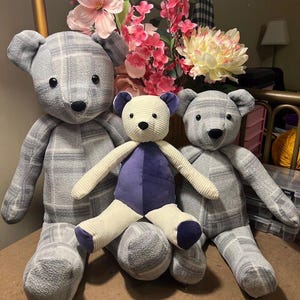 Puede incluir: Tres osos de peluche est&aacute;n dispuestos juntos. Dos osos m&aacute;s grandes est&aacute;n hechos de una tela polar a cuadros gris y blanca con ojos y narices de bot&oacute;n negros. Un oso m&aacute;s peque&ntilde;o en el centro tiene extremidades de pana crema y torso morado.