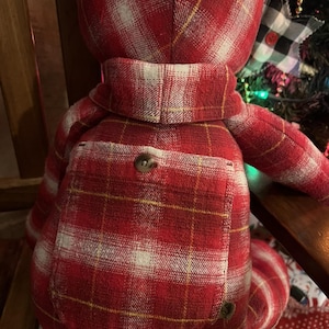 Puede incluir: Un oso de peluche confeccionado con tela de cuadros rojos, blancos y amarillos. El oso presenta un bolsillo cuadrado prominente en la espalda asegurado con un bot&oacute;n marr&oacute;n, un detalle tipo cuello alrededor del cuello y orejas redondeadas, todo igual.