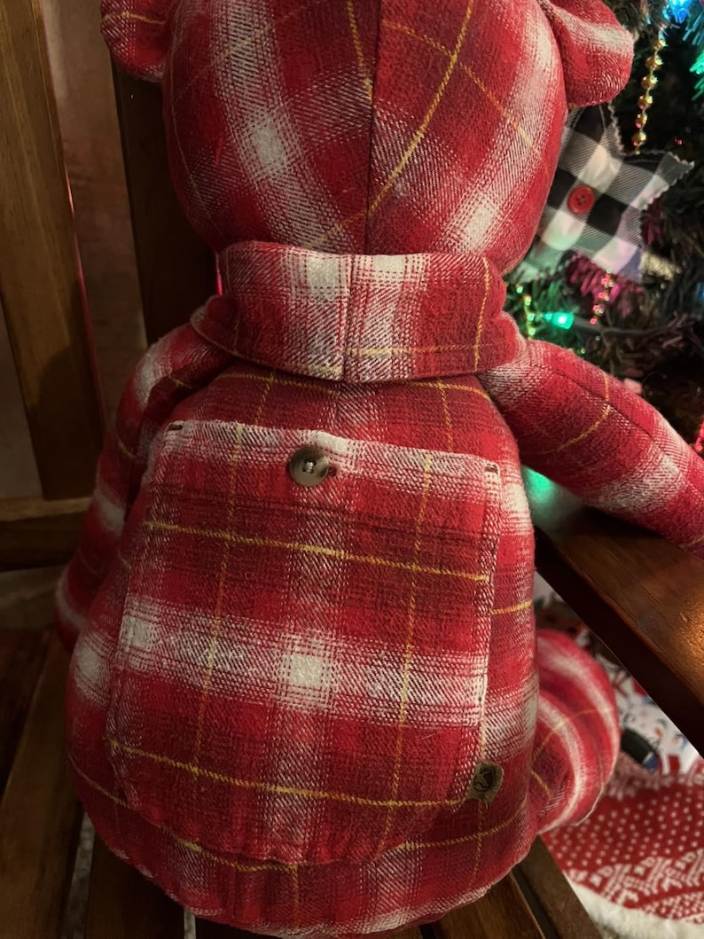 Puede incluir: Un oso de peluche confeccionado con tela de cuadros rojos, blancos y amarillos. El oso presenta un bolsillo cuadrado prominente en la espalda asegurado con un bot&oacute;n marr&oacute;n, un detalle tipo cuello alrededor del cuello y orejas redondeadas, todo igual.