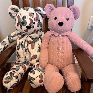 Puede incluir: Dos osos de peluche sentados uno al lado del otro. El oso de la izquierda presenta una tela blanca con un patr&oacute;n de mariposas coloridas, mientras que el de la derecha es de tela malva s&oacute;lida con textura de puntos, cuello de encaje y tres botones.