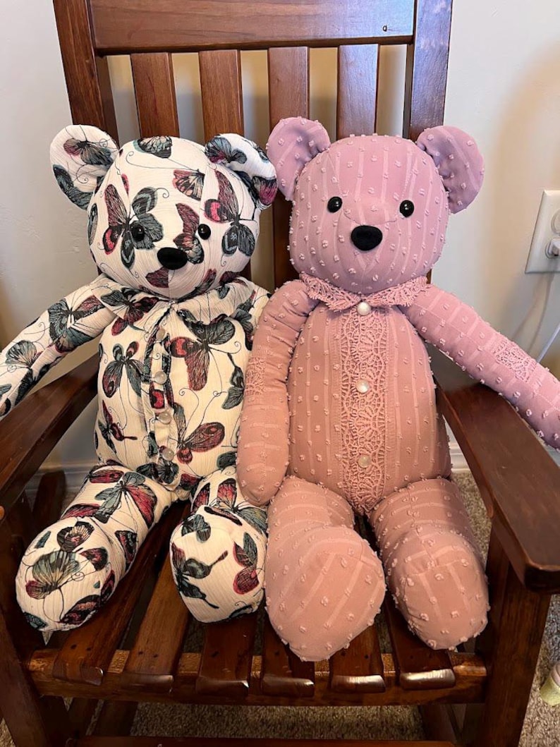 Puede incluir: Dos osos de peluche sentados uno al lado del otro. El oso de la izquierda presenta una tela blanca con un patr&oacute;n de mariposas coloridas, mientras que el de la derecha es de tela malva s&oacute;lida con textura de puntos, cuello de encaje y tres botones.