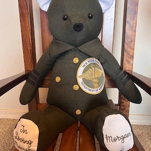 Puede incluir: Un oso de peluche de tela verde oliva oscuro con una chaqueta de uniforme con botones dorados y un parche del New York City Department of Sanitation. Tiene orejas interiores y plantas de los pies blancas con el texto In Loving Memory y Morgan.