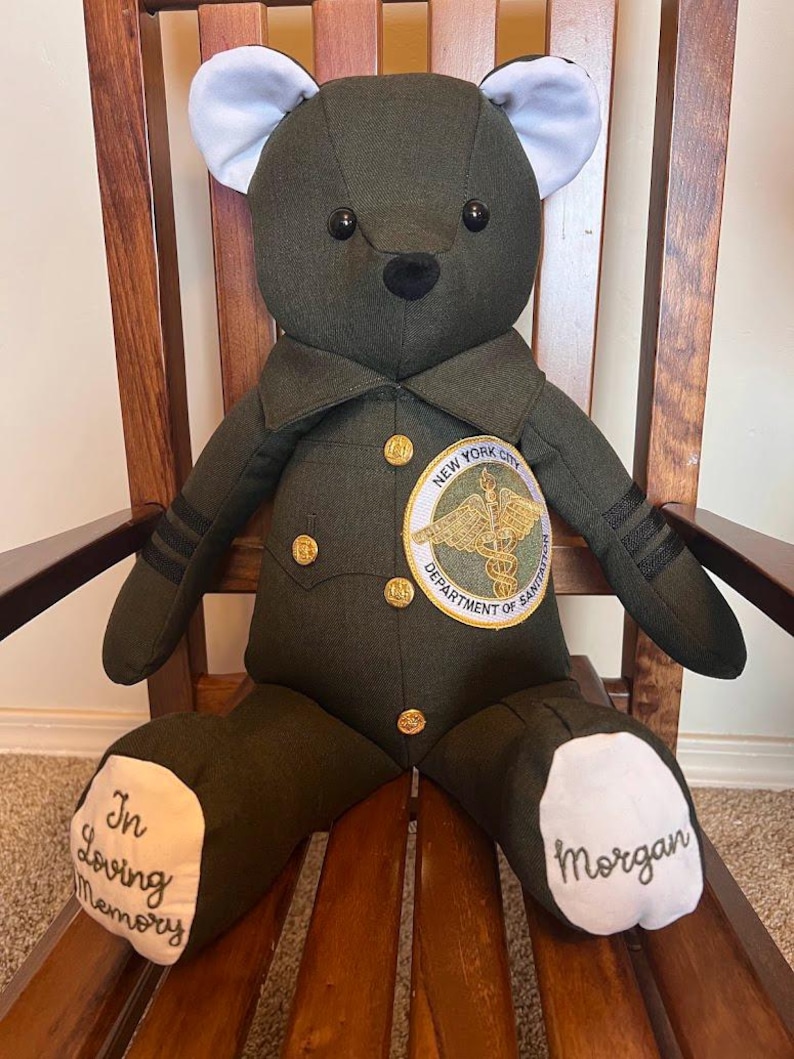Puede incluir: Un oso de peluche de tela verde oliva oscuro con una chaqueta de uniforme con botones dorados y un parche del New York City Department of Sanitation. Tiene orejas interiores y plantas de los pies blancas con el texto In Loving Memory y Morgan.