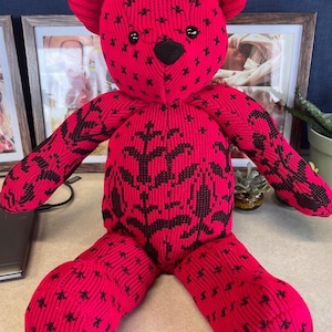 Puede incluir: Un oso de peluche hecho de tela acanalada de color rosa brillante con un patr&oacute;n floral y de estrellas en negro. El oso presenta ojos de pl&aacute;stico negro, una nariz negra y la palabra "Mom" bordada en cursiva blanca en la planta del pie derecho.