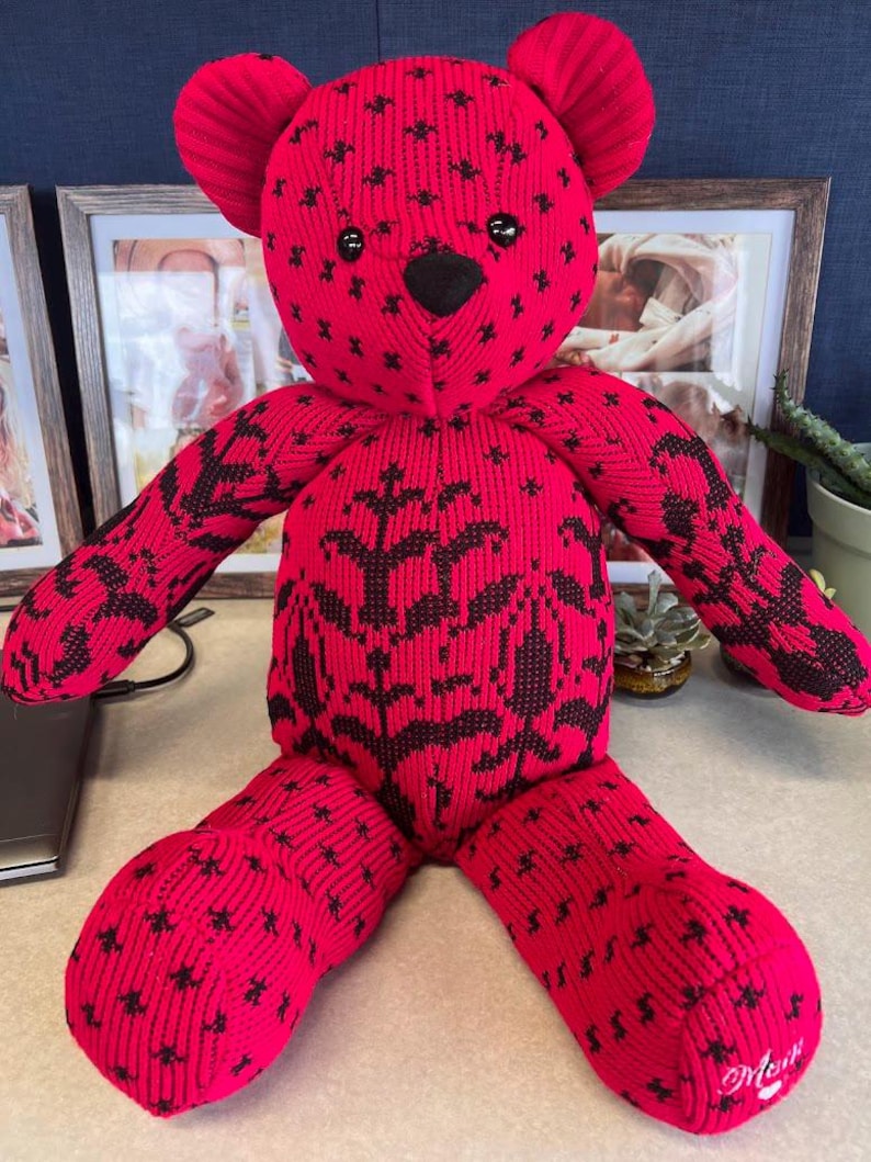 Puede incluir: Un oso de peluche hecho de tela acanalada de color rosa brillante con un patr&oacute;n floral y de estrellas en negro. El oso presenta ojos de pl&aacute;stico negro, una nariz negra y la palabra "Mom" bordada en cursiva blanca en la planta del pie derecho.