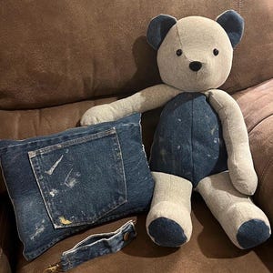 Puede incluir: Un oso de peluche hecho de tela gris claro con detalles de mezclilla azul oscuro en las orejas, el torso y los pies. Junto a &eacute;l hay un coj&iacute;n rectangular de mezclilla con un bolsillo con salpicaduras de pintura y una correa a juego.