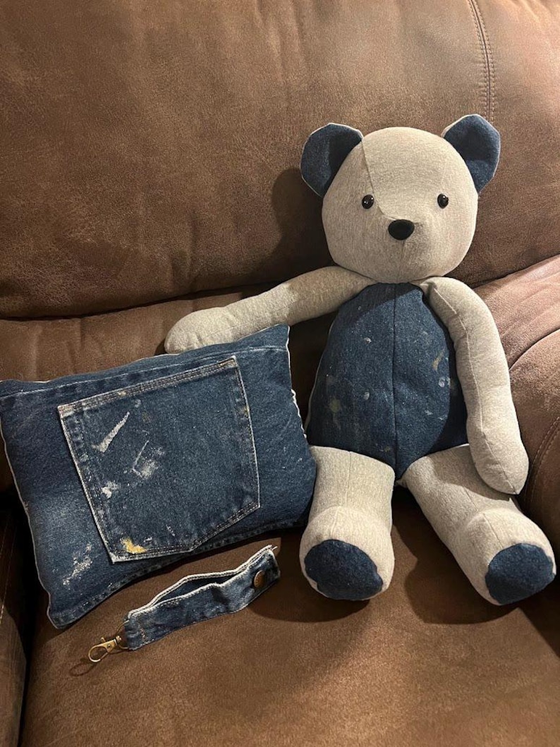 Puede incluir: Un oso de peluche hecho de tela gris claro con detalles de mezclilla azul oscuro en las orejas, el torso y los pies. Junto a &eacute;l hay un coj&iacute;n rectangular de mezclilla con un bolsillo con salpicaduras de pintura y una correa a juego.