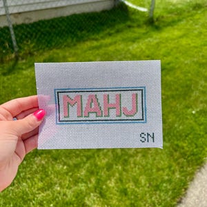 以下が含まれることがあります： ピンクと緑の文字で「MAHU」と刺繍されたニードルポイントキャンバス。青と黒のボーダーで囲まれています。イニシャル「SN」は右下にあります。キャンバスは緑の草を背景に保持されています。