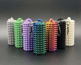 May include: A collection of seven cylindrical keychains in various colors, including silver, green, purple, blue, rainbow, black, and white. Each keychain features a textured, spiky surface and a metal clasp for attaching keys.