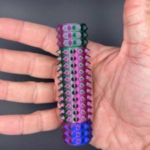 May include: A multi-colored, cylindrical object with a textured surface. The object features a gradient of colors, including purple, green, and blue. The design incorporates a series of conical spikes around the body. The object is held in a hand.