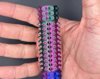 May include: A multi-colored, cylindrical object with a textured surface. The object features a gradient of colors, including purple, green, and blue. The design incorporates a series of conical spikes around the body. The object is held in a hand.