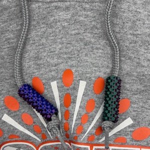 May include: Close-up of a gray hoodie with a drawstring. The hoodie features a graphic design with orange and white accents. The drawstring ends have blue and green textured details.