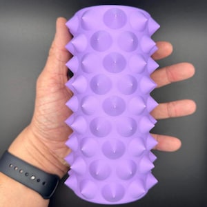 Puede incluir: Rodillo de espuma morado con una superficie texturizada cubierta de pequeñas púas. El rodillo está diseñado para el automasaje y la recuperación muscular.
