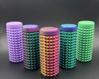 Könnte beinhalten: Fünf zylindrische Behälter mit strukturierten, stacheligen Oberflächen und flachen, glitzernden Deckeln. Die Behälter sind in den Farbtönen Lila, Grün, Gold und Blau gehalten. Die Behälter sind in einer Reihe vor einem dunklen Hintergrund angeordnet.
