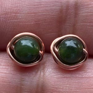 May include: A pair of stud earrings featuring a round, dark green gemstone set in a rose gold-colored metal. The metal is intricately wrapped around the gemstone, creating a delicate, yet secure setting. The earrings are designed for pierced ears.
