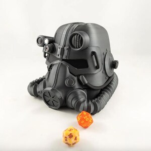 Puede incluir: Un casco de armadura de potencia Fallout impreso en 3D de color negro y gris con una máscara de gas y un par de dados naranja y amarillo.