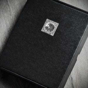 May include: A black box with a circular sticker on the top. The sticker features a detailed, grayscale design with a bird and other symbols. The box is on a wooden surface.