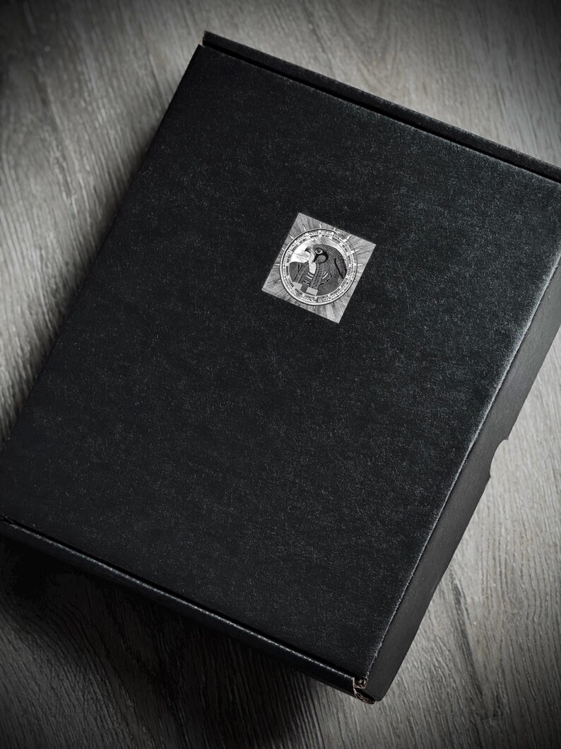 May include: A black box with a circular sticker on the top. The sticker features a detailed, grayscale design with a bird and other symbols. The box is on a wooden surface.