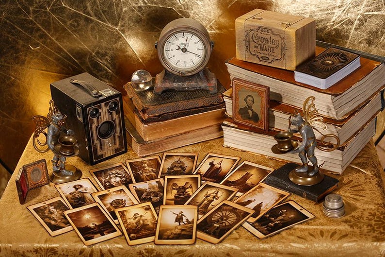 May include: A vintage camera, a clock, a stack of books, and a small figurine are arranged on a table with a gold-colored tablecloth. The books have a worn, antique look and are stacked on top of each other. The camera is a black and brown box camera with a lens on the front. The clock is a round, brass clock with Roman numerals. The figurine is a small, gold-colored angel with wings. The table is covered in a gold-colored tablecloth with a pattern of small, white dots.