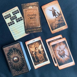 May include: A tarot deck with a gold and black design on the back of the cards. The cards are laid out on a dark surface. The cards include the Queen of Wands, Eight of Swords, and The Tower. The deck is titled "Crowley and Waite Victorian Tarot".