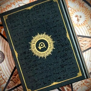 K&ouml;nnte beinhalten: Ein dunkelgr&uuml;nes Buch mit goldenen Akzenten und einem Sonnenstrahl-Design. Das Cover zeigt ein goldenes Sonnenstrahl-Emblem mit einem Auge in der Mitte und goldfarbenen Zierleisten. Das Buch befindet sich vor einem Hintergrund mit Himmelsbildern.
