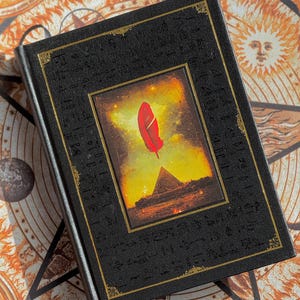 K&ouml;nnte beinhalten: Ein schwarzes Hardcover-Buch mit goldenen Verzierungen und einem zentralen Bild einer roten Feder &uuml;ber einer Pyramide. Der Einband hat gepr&auml;gte Symbole im Hieroglyphenstil. Der Hintergrund zeigt ein himmlisches Design.