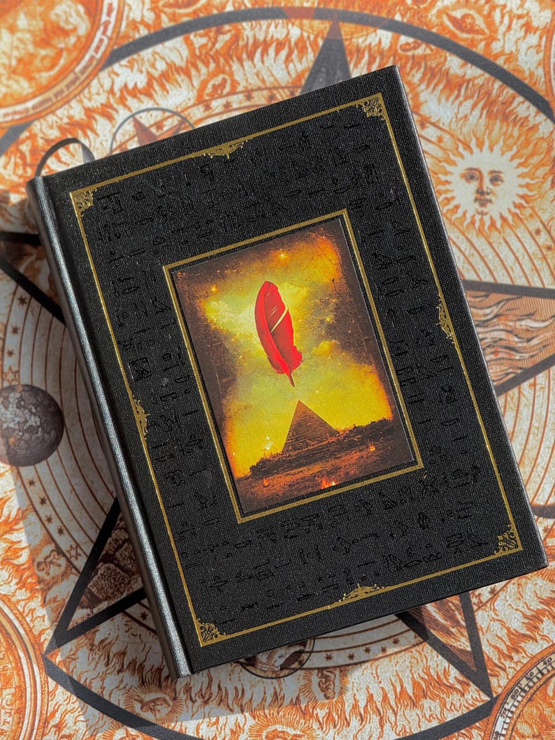 K&ouml;nnte beinhalten: Ein schwarzes Hardcover-Buch mit goldenen Verzierungen und einem zentralen Bild einer roten Feder &uuml;ber einer Pyramide. Der Einband hat gepr&auml;gte Symbole im Hieroglyphenstil. Der Hintergrund zeigt ein himmlisches Design.